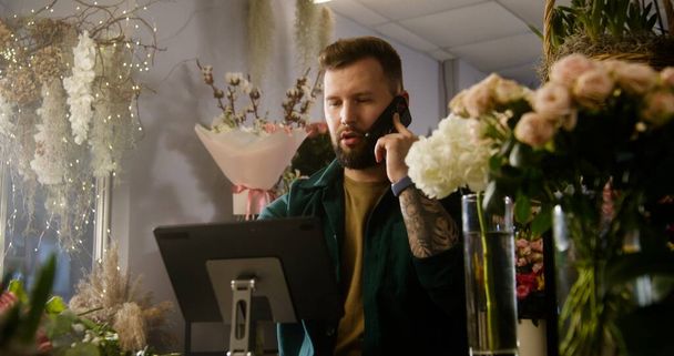 Férfi vállalkozó, tulajdonos, virágárus beszél telefonon, és veszi az ügyfél érdekében online digitális tabletta. Virágok a vázákban a virágüzletben. Virágzás, virágos kisvállalkozások és vállalkozói szellem. - Fotó, kép