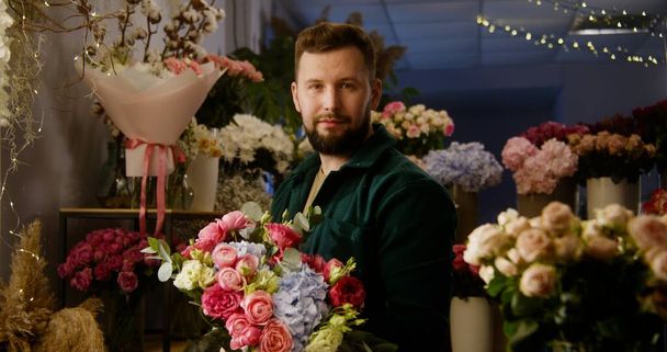 Professional florist collects and keeps beautiful bouquet in hands, smiles and looks at camera. Vases with fresh flowers stand behind man. Retail floral business and entrepreneurship concept. Portrait - Foto, Bild