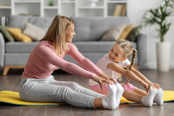 Happy Young Mother And Little Daughter Doing Sports Together At Home, Smiling Mom And Cute Female Child Reaching Toes With Hand While Sitting On Fitness Mat, Stretching Legs And Arms, Closeup - 写真・画像