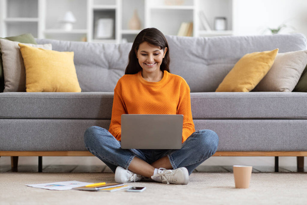 Lächelnde, ziemlich langhaarige Millennial Lady in lässigem Outfit sitzt auf dem Boden neben der Couch im Wohnzimmer, Projektmanager arbeitet von zu Hause aus online, benutzt Laptop, trinkt Kaffee, kopiert Raum - Foto, Bild