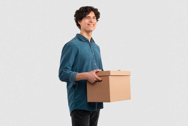 Koncept dodávky. Veselý Evropan nesoucí kartónovou krabici stojící na pozadí Bílého studia, dívající se stranou. Mladý muž držící balík Reklama Velká nabídka dodávky - Fotografie, Obrázek