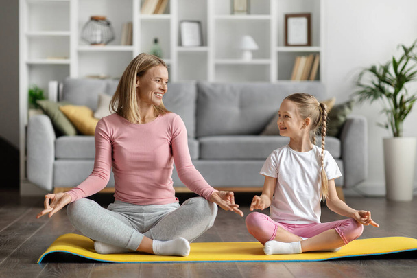 Glückliche Mutter und süße kleine Tochter meditieren gemeinsam zu Hause, sportliche Mutter und Kind sitzen auf der Fitnessmatte in Lotus-Pose, praktizieren Yoga oder Pilates, genießen gesunden Lebensstil, kopieren Raum - Foto, Bild