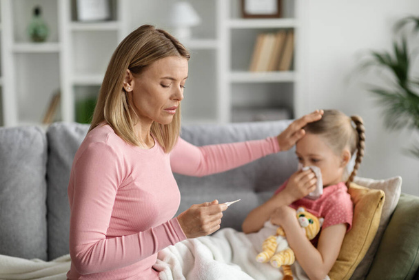 Seasonal Flu Concept. Worried Mother Taking Care Of Her Ill Daughter At Home, Caring Mom Touching Kids Forehead And Looking At Thermometer, Checking Temperature Of Sich Child, Closeup Shot - Foto, immagini