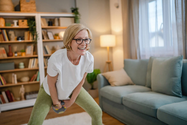 Una mujer rubia caucásica madura entrenando con kettlebell girya campana rusa en casa ejercicio femenino en su apartamento concepto de estilo de vida saludable - Foto, imagen