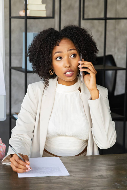 Women entrepreneurship. Vertical portrait of successful pretty young black businesswoman in formal outfit sitting at desk, signing papers, talking on phone with business partner or client - Фото, изображение