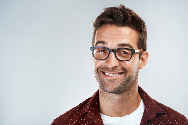 One of those cheeky smiles. Portrait of a cheerful young man wearing glasses and smiling brightly while standing against a grey background - Foto, Imagem