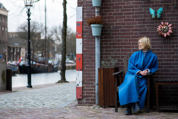 An attractive adult woman in a blue coat looks to the side. Behind her is a brick wall. Cool weather. Travel and adventure. - Foto, imagen