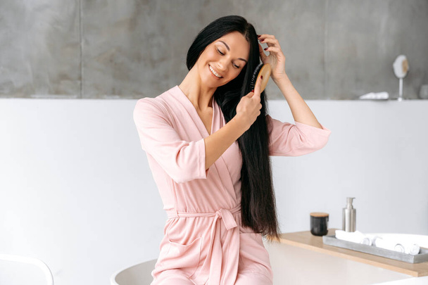 Female beauty routine. Combing hair. Beautiful caucasian brunette woman, sits in a stylish modern bathroom, in a bathrobe, combs her long hair with a wooden comb, smiles - Photo, Image