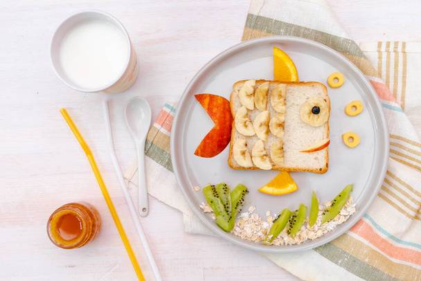 Funny cute fish shape sandwich toast bread with banana, apple,orange, milk,honey. Kids childrens baby's sweet dessert healthy breakfast lunch food art on plate,wooden background close up,top view. - Photo, Image