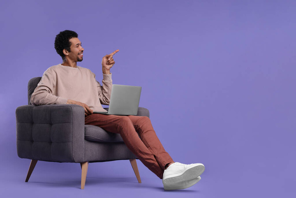 Happy man with laptop sitting in armchair and pointing at something on purple background. Space for text - Zdjęcie, obraz