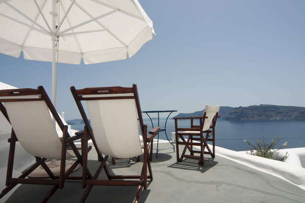 Relájese con estilo en la terraza de la azotea con impresionantes vistas de la caldera de Santorini y el mar Egeo, con comodidad bajo un paraguas en cómodos sillones. Sus vacaciones de verano perfectas. - Foto, Imagen