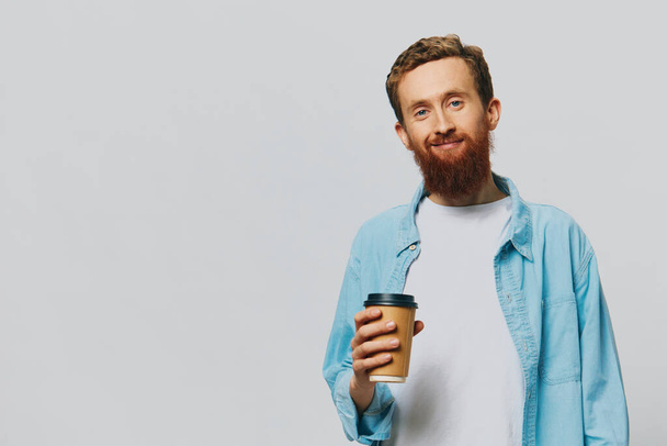 Man hipster met een kopje koffie met een glimlach op een grijze achtergrond in een blauw shirt en wit t-shirt. Hoge kwaliteit foto - Foto, afbeelding