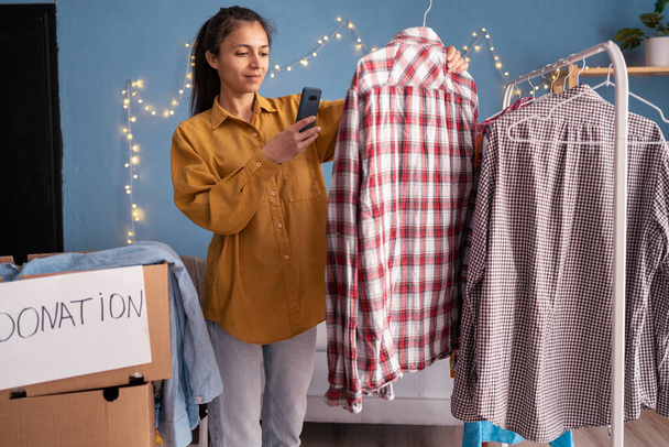 Ділова жінка вибирає одяг для пожертвування, фотографує сорочки за допомогою смартфона перед відправкою. Концепція пожертвування або переробки. Копіювати простір
 - Фото, зображення