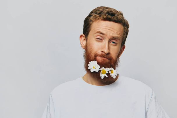 Портрет смішного чоловіка в білій футболці з квітами ромашки в бороді на білому ізольованому тлі, місце для тексту. Святкова концепція та привітання. Високоякісна фотографія - Фото, зображення