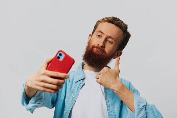 Мужчина-хипстер с телефоном в руке блоггер улыбается на сером фоне в голубой рубашке и белой футболке, разговаривает по телефону и селфи. Высокое качество фото - Фото, изображение