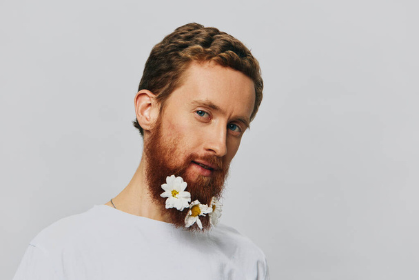 Portrait of a funny man in a white T-shirt with flowers daisies in his beard on a white isolated background, copy place. Holiday concept and congratulations. High quality photo - Photo, Image