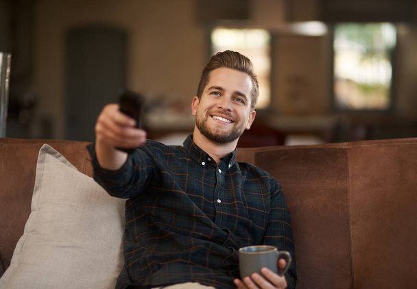 Relax, coffee and a man watching tv on a sofa in the living room of his home with a remote for streaming. Smile, tea and a happy young male person using a subscription service on his television. - Foto, Imagen