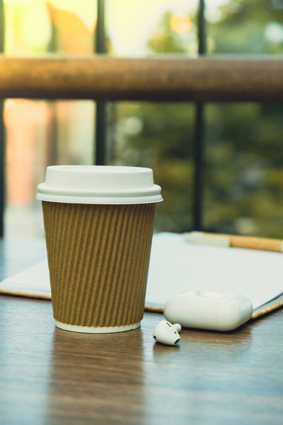 Warme latte koffie in ambachtelijke recycling papieren beker met papieren notebook met draadloze hoofdtelefoon. Een afhaalbekertje op de cafe tafel. Freelance werkruimte notebook met koffie voor productief werk en studie - Foto, afbeelding