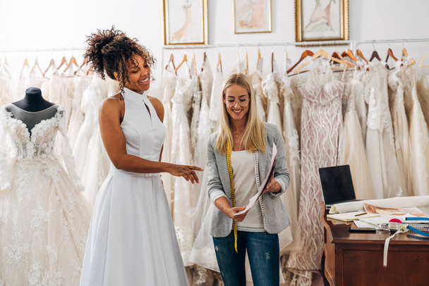 A beautiful bride and a tailor discuss together sketches for a wedding dress. - Foto, afbeelding