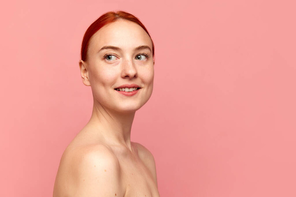 Close up shot of beautiful girl with red hair and bare shoulders looking away and smiling over pink studio background. Model with perfect skin. Beauty, spa, cosmetology, skin care, emotion concept - Foto, Bild