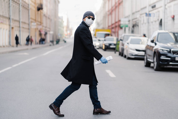 ウイルスは-19をcovid 。男は黒いコート、帽子、サングラスを身に着けている道路を横断し、パンデミックウイルス保護のための保護マスクを着用し、多くの輸送と人々と忙しい通りで屋外をポーズします - 写真・画像