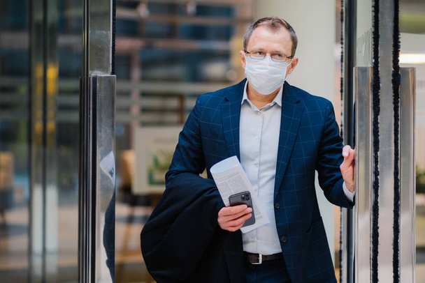 Covid-19, pandemický koronavirový koncept. Pracovník z kanceláře opouští práci, nosí lékařskou masku na obličej pro infekční onemocnění šíří, obléká se formálně, drží noviny a smartphone. Koncepce ochrany zdraví a bezpečnosti - Fotografie, Obrázek