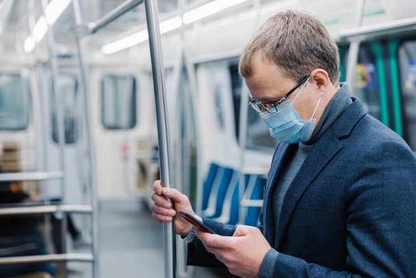 El hombre serio viaja para trabajar en metro lleva máscara protectora de virus siempre en contacto con las poses celulares modernas en metro vacío o metro. Concepto de distancia. Transporte público durante coronavirus
 - Foto, Imagen