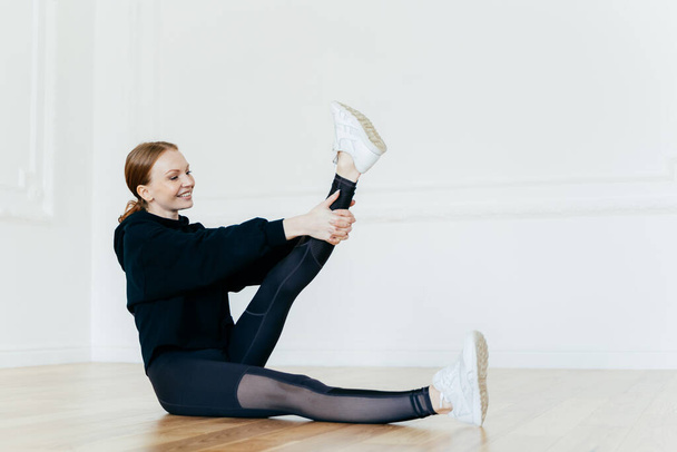 Athletic woman stretches leg, has satisfied facial expression, wears black clothes, white sportshoes, sits on floor, does pilates exercises, has joyful look, exercises acitively. Sport concept - Foto, Bild