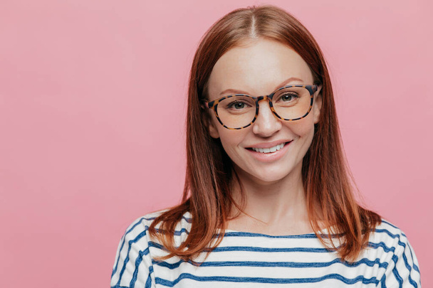 Close up shot of adorable young female student wears optical glasses, has tender smile, rejoices holidays wears striped clothes, models over pink background with copy space for your information. - Photo, Image