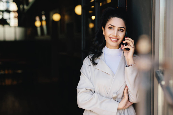Elegant brunette businesswoman with dark eyes and full lips dressed in white clothes communicating over telephone having charming smile looking up with thoughtful expression. Technology concept - Photo, Image