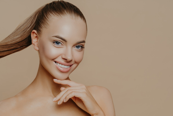 Tranquil undressed woman with healthy perfect skin, dark hair combed in pony tail, enjoys facial treatments, has well cared complexion, stands against beige studio wall, empty space. Skin care concept - Photo, Image