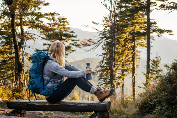 Frau mit Rucksack und Thermoskanne ruht auf Bank im Wald. Entspannung beim Wandern in den Bergen - Foto, Bild