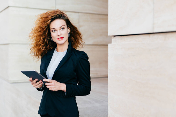 Beautiful businesswoman with curly fluffy hair, wearing elegant clothes, holding tablet in hands, waiting for her business partner near cafe, using free internet conncection. People, career, work - Foto, imagen