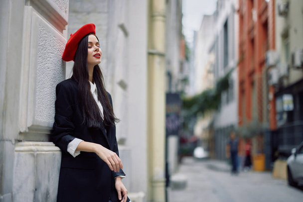 Woman standing near a wall in the city wearing a stylish jacket and red beret with red lips, travel and leisure, French style of dress, spring. High quality photo - Photo, Image