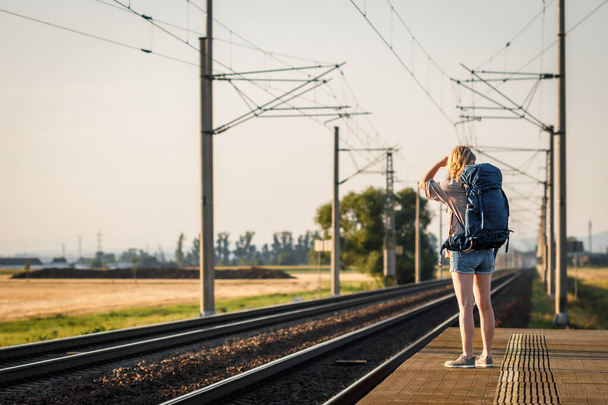 Woman traveler with backpack waiting for train at railway station. Female tourist traveling at summer vacation alone - Foto, Imagen