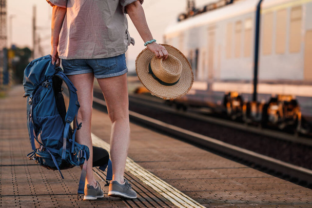 Ταξιδέψτε με τρένο στις καλοκαιρινές διακοπές. Τουριστική με σακίδιο και καπέλο περιμένει στο σιδηροδρομικό σταθμό - Φωτογραφία, εικόνα