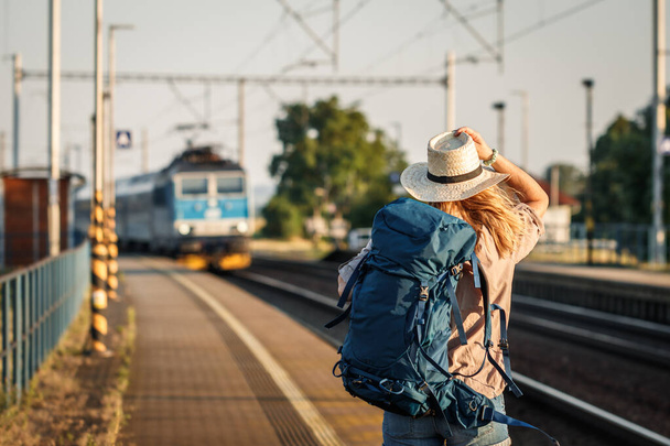 Female backpacker traveling by train alone. Woman with backpack and straw hat standing at railroad station platform and looking at arriving train - Foto, Imagem