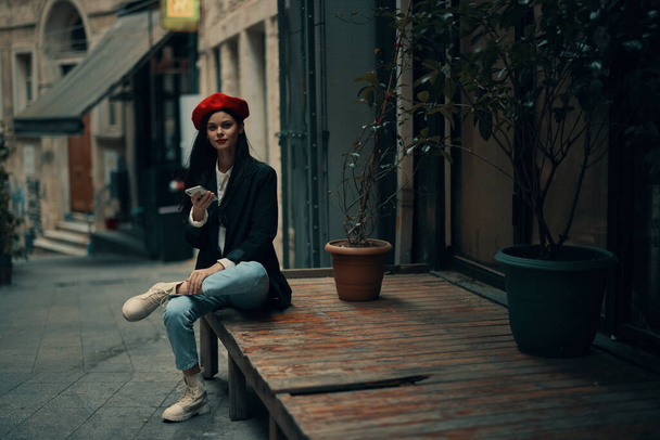 Stylowa kobieta turysta w stylowych ubraniach w kurtce i czerwony beret siedzi na zewnątrz z telefonem w ręku, telefon komórkowy w podróży, francuski styl, kolor kinowy. Wysokiej jakości zdjęcie - Zdjęcie, obraz