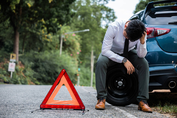 若い実業家と1人の成熟した男は、警告赤の三角形の看板を背景に壊れた車に座っている。成熟した男が心配している間にビジネスマンは車輪を変えています. - 写真・画像