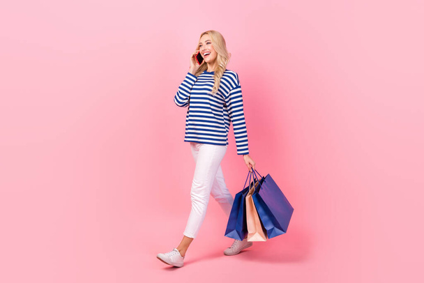 Full length photo of cute cheerful woman dressed striped shirt holding shoppers communicating modern device isolated pink color background. - Foto, afbeelding