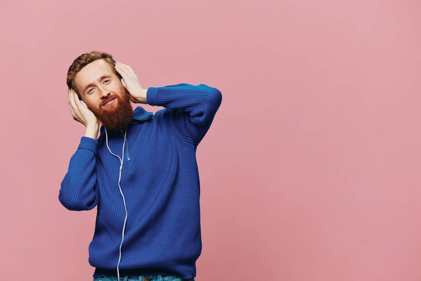 Egy vörös hajú férfi portréja, aki fejhallgatót visel, mosolyog és táncol, rózsaszín háttérrel zenét hallgat. Egy szakállas hipszter. Kiváló minőségű fénykép - Fotó, kép