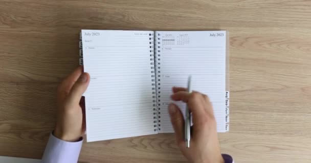 Man flips pages of diary calendar holding pen in hand. Male manager ready to write working schedule sitting at wooden table in office - Footage, Video
