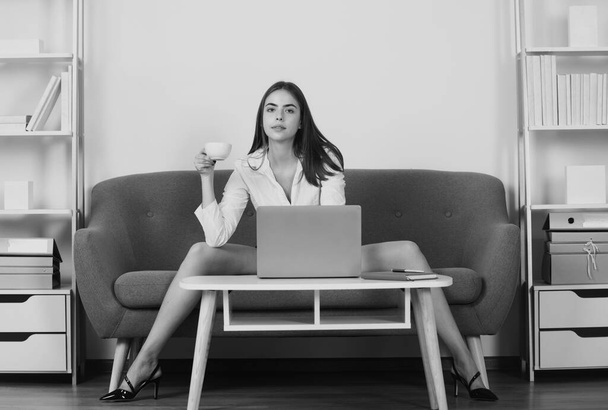 Sexy businesswoman with cup coffee and sexy legs. Seductive woman, sensual secretary sit on sofa in office. Sexy businesswoman with laptop computer wearing white suit with decollete sitting on sofa - Фото, зображення