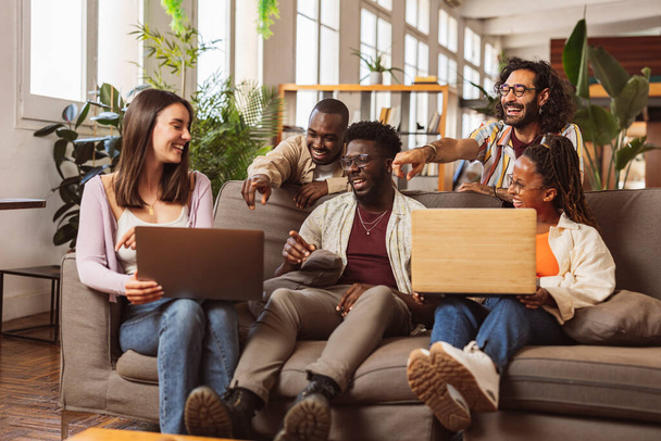 Group of happy multiethnic coworkers in casual clothes relaxing on a sofa in a modern office room and using laptop during the day. Friends in apartment, university students end-of-year project - Фото, изображение