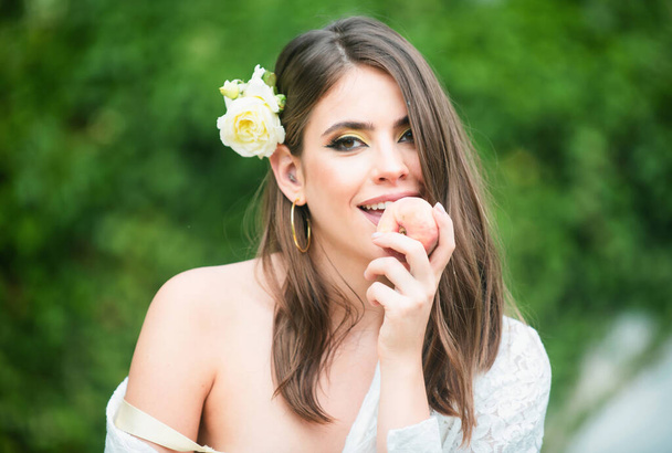 Sexy girl on summer picnic on a grass in the park. Outdoor fashion photo of beautiful young woman with rose in hair eating peach - Foto, imagen
