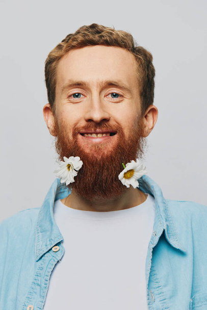 Retrato de um homem engraçado em uma camiseta branca com flores margaridas em sua barba em um fundo isolado branco, lugar de cópia. Conceito de férias e parabéns. Foto de alta qualidade - Foto, Imagem