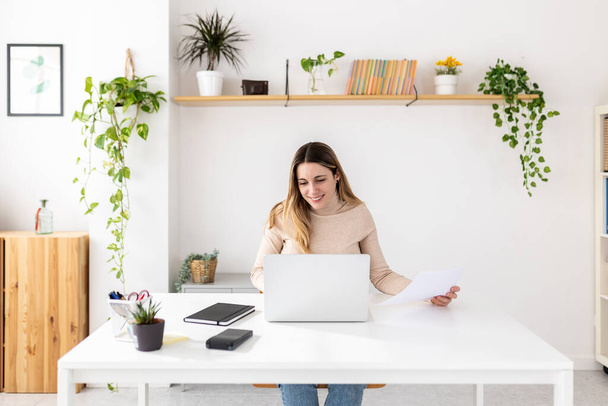 Młoda, dorosła kobieta pracująca na laptopie, siedząca na biurku w biurze. Millennial blondynka bizneswoman patrząc na ekranie komputera podczas sprawdzania dokumentów.  - Zdjęcie, obraz