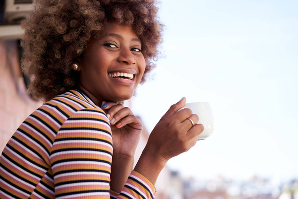 Šťastná mladá černoška s afro vlasy pózuje při pohledu na fotoaparát s úsměvem. Veselá Afroameričanka drží šálek kávy venku. Portrét ženy s pozitivním výrazem. Kopírovat prostor. - Fotografie, Obrázek