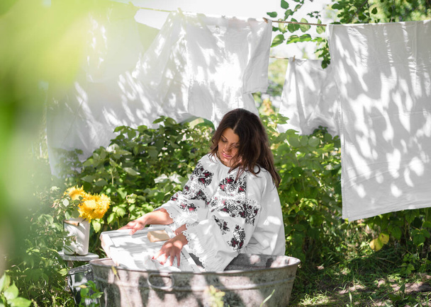 Ένα κορίτσι ισπανόφωνης εμφάνισης είναι ντυμένο με ένα Ουκρανικό φόρεμα Vyshyvanka στον κήπο. Το κορίτσι πλένει και κρεμάει τα πλυμένα ρούχα στη γραμμή ρούχων στον κήπο. Διακοπές ουκρανική vyshyvanka έννοια ημέρα - Φωτογραφία, εικόνα