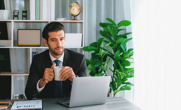 Nowoczesny profesjonalny biznesmen w nowoczesnym biurze przy użyciu laptopa do pracy z kawą w ręku. Staranny pracownik biurowy pracujący nad notatnikiem komputerowym w swoim biurze. żarliwy - Zdjęcie, obraz